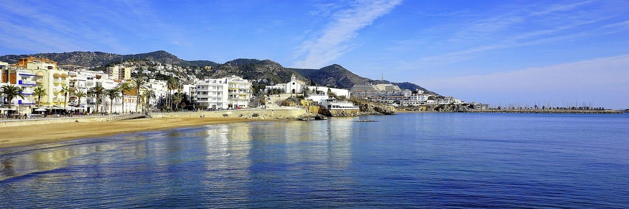 The beach at Sitges curving away to a low headland, white buildings along the front and hills rising behind the town under a clear sky.