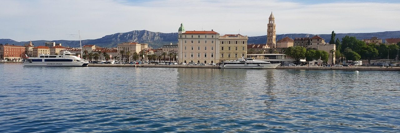 The waterfront at Split seen from across the harbour: ferries moored along the Riva, the long stone frontage of Diocletian's Palace behind a line of palms, the cathedral bell tower above it and bare grey hills on the skyline.