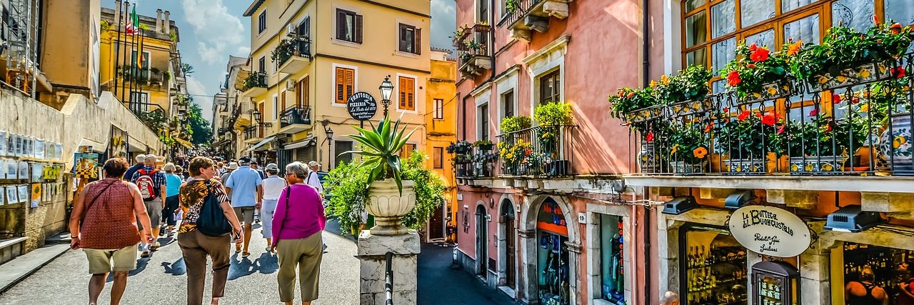 Corso Umberto in Taormina crowded with people between shopfronts, balconies hung with flowers above them and awnings shading the street.