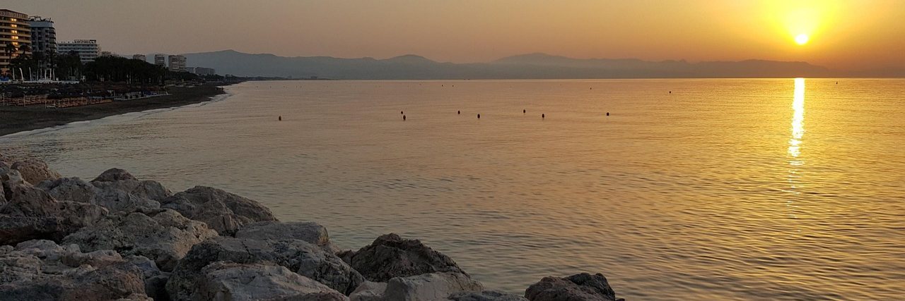 A long bay at sunrise, boulders in the foreground and apartment blocks lining the beach away to the left, with mountains across the water.