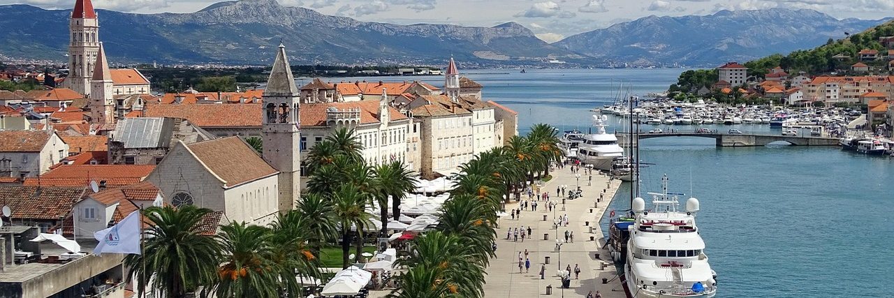 Trogir from above: terracotta roofs and two church towers on the left, the palm-lined waterfront promenade with yachts moored along it, and the channel and mainland mountains beyond.