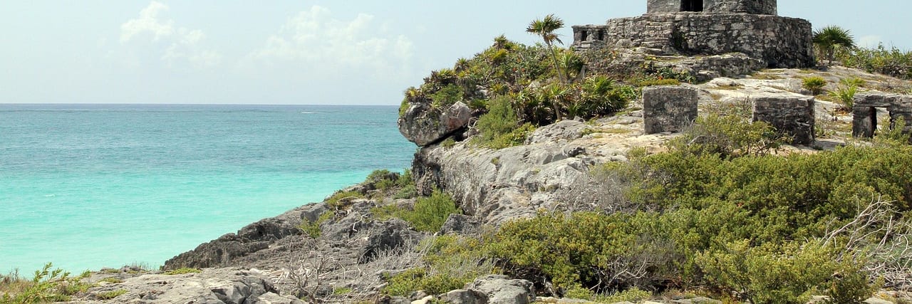A small stone Mayan temple on a low cliff above turquoise sea at Tulum, scrub and palms around it.